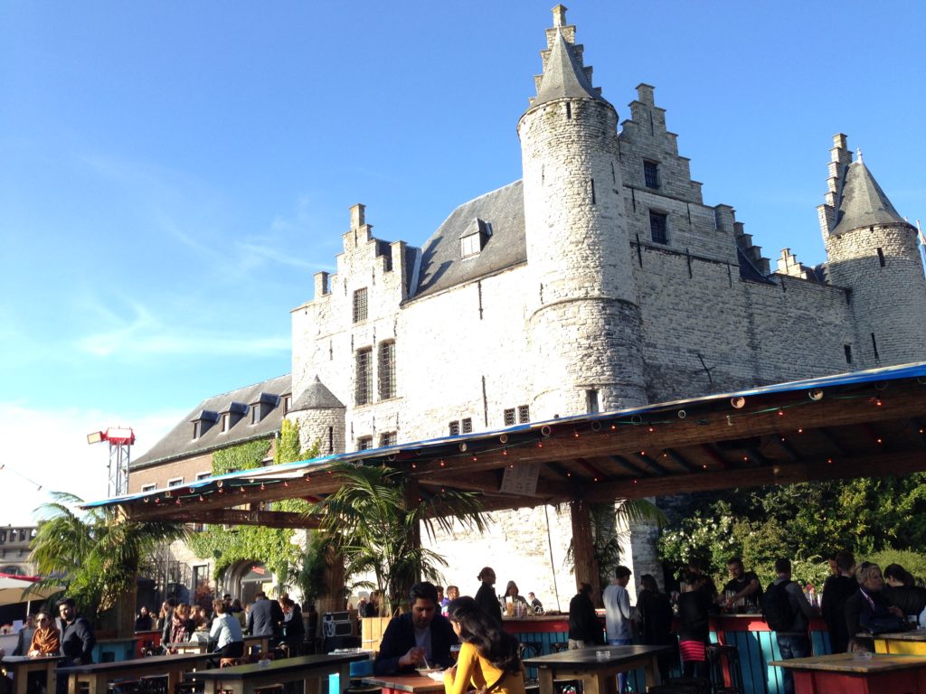 people eating and drinking at tables on a patio below a castle