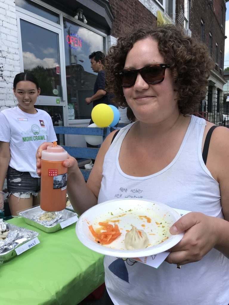 woman holding hot sauce in one hand and plate with momo in other hand while volunteer server looks on at Loga's Corner in Toronto